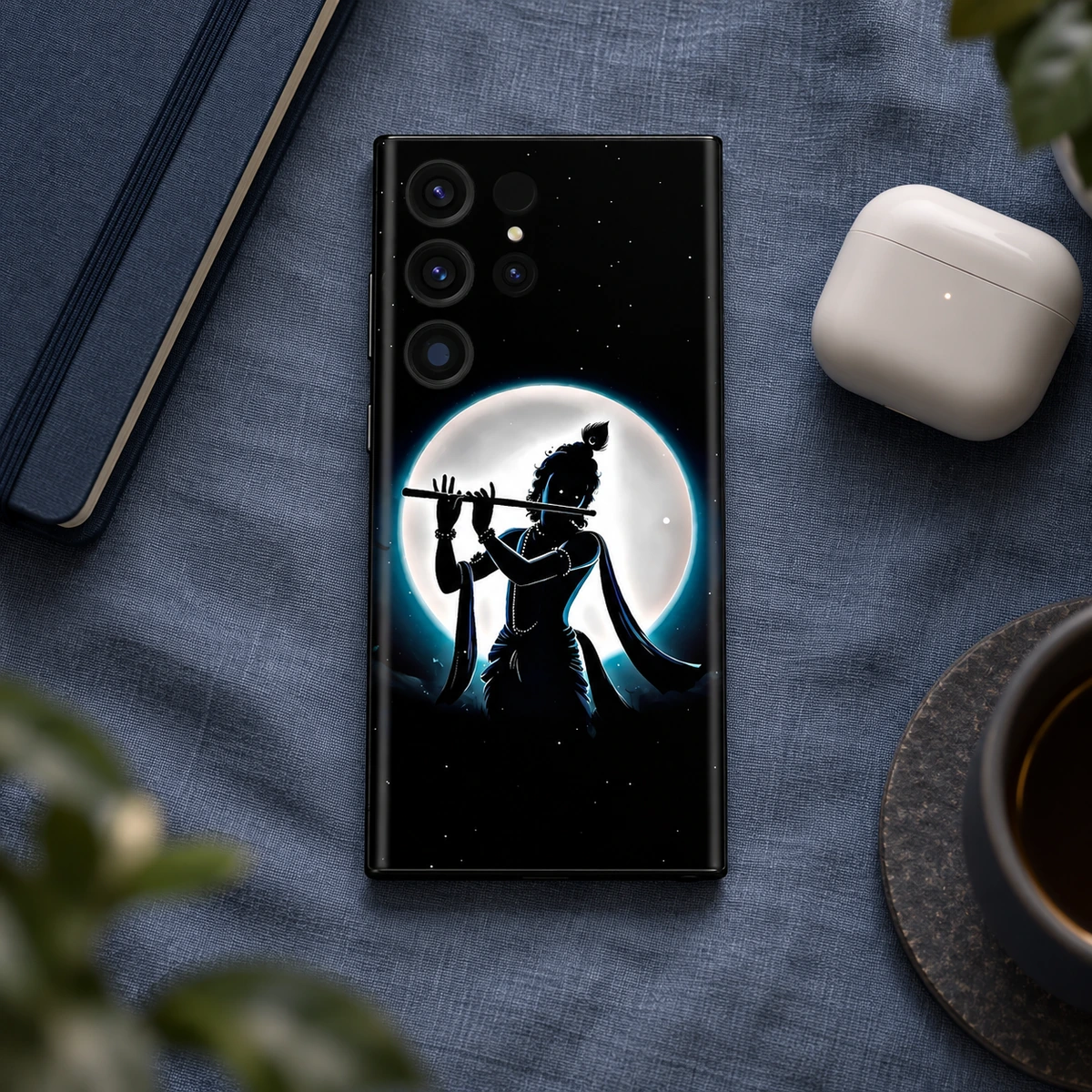 Jay Shree Krishna Full Moon Flute Mobile Skin - Night desk flatlay Black Krishna full moon mobile skin on blue linen beside notebook and earbuds case