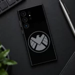 Black Marvel Shield mobile skin with grey circular eagle emblem, tech desk flat lay