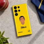 Yellow Dhoni Thala mobile skin lying on pale concrete beside a cricket ball and navy fabric