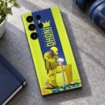 Yellow Dhoni photo mobile skin with crown, vertical MAHENDRA DHONI lettering and kneeling cricketer, modern desk