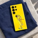 Yellow MS Dhoni celebration mobile skin showing two cricket players embracing, navy fabric lifestyle
