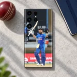 Virat Kohli mobile skin showing a raised bat and helmet celebration in blue India uniform, cricket desk flat lay