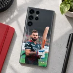 Virat Kohli photo mobile skin showing bat portrait in black-and-red cricket jersey, concrete desk flat lay