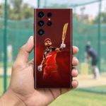 Virat Kohli mobile skin with raised bat and red cricket kit against burgundy, practice-net hand-held view